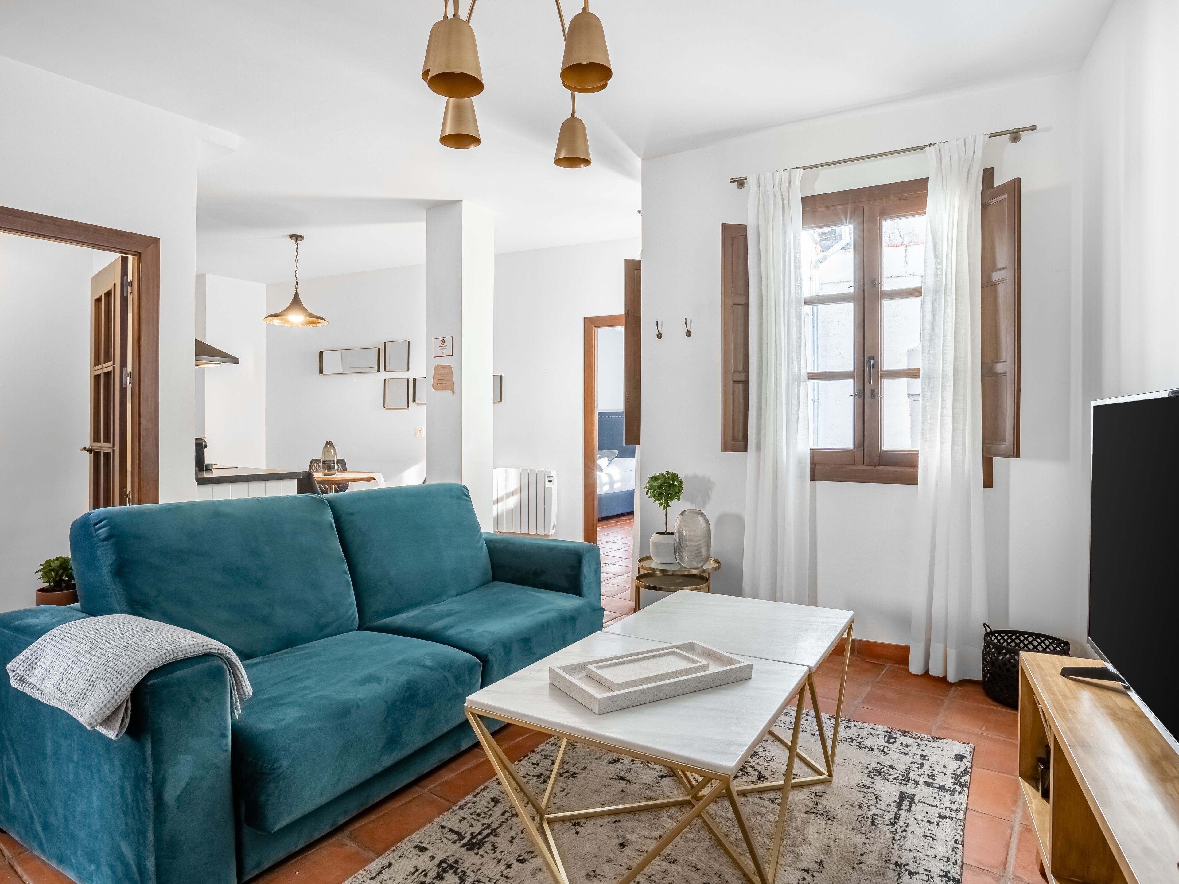 A modern living room with a teal sofa, a white coffee table, and a TV. There are wooden doors, a window with curtains, and a small table with a plant.