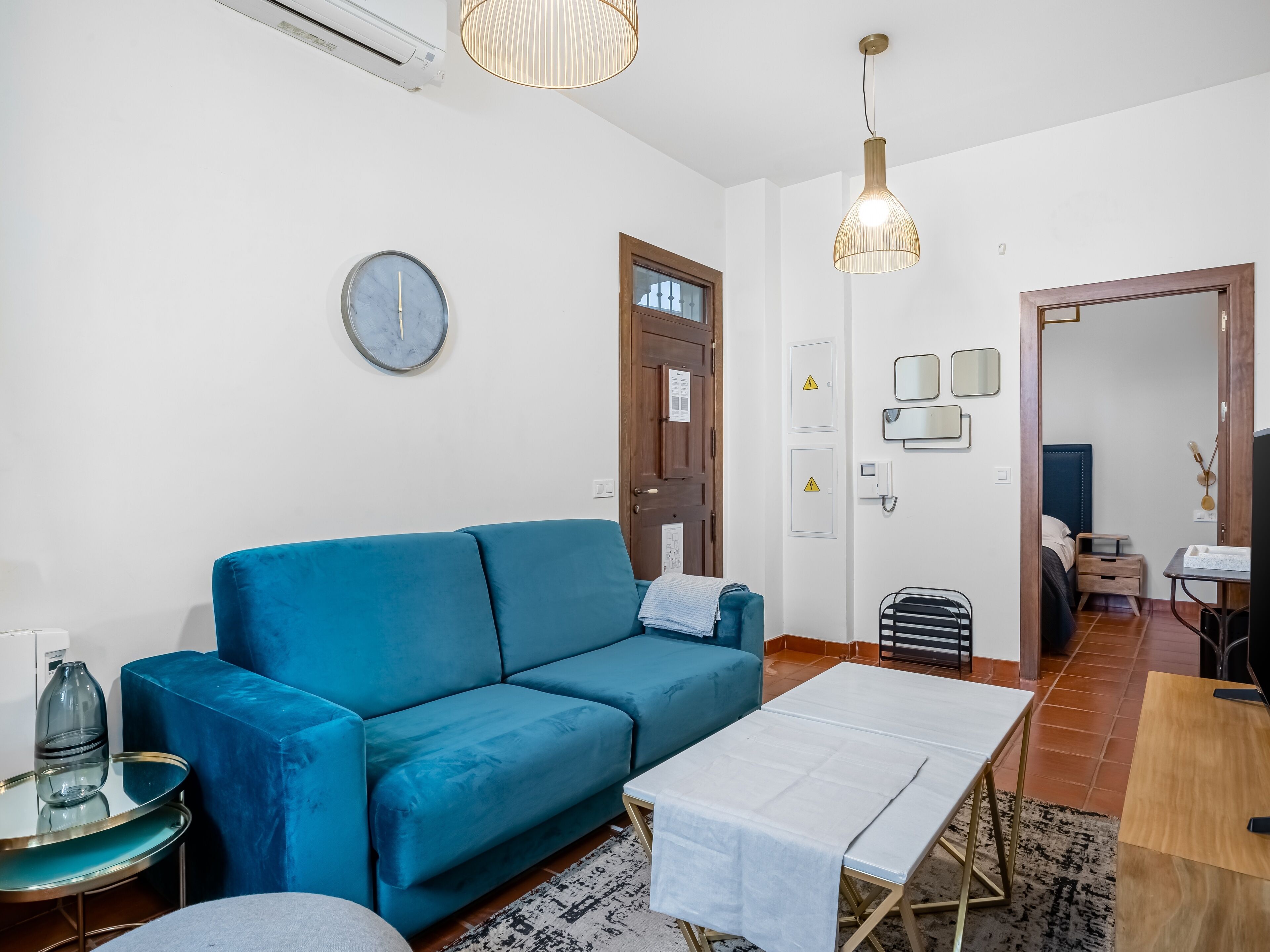 A modern living room with a blue sofa, a white coffee table, and a dining table. There is a clock on the wall and a door with a wooden frame.