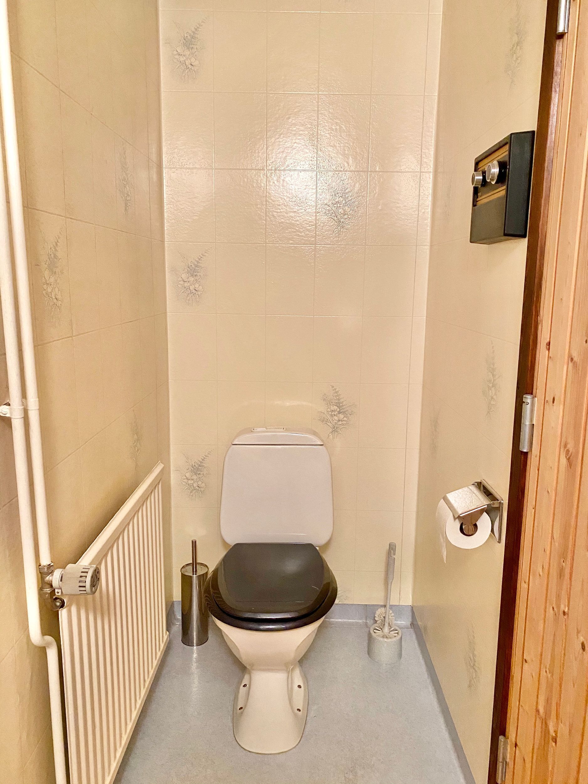 Une petite salle de bain étroite, équipée d’un WC, d’un radiateur et d’un porte-rouleau de papier toilette.