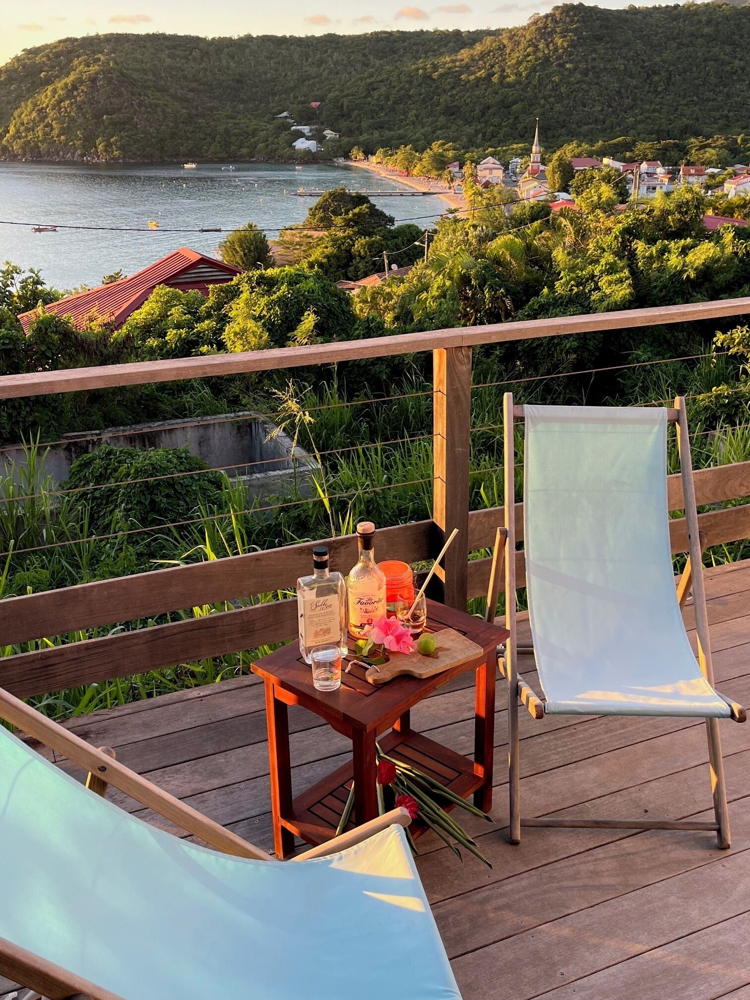A wooden deck with a table set for two, overlooking a coastal area with a beach and a hill.