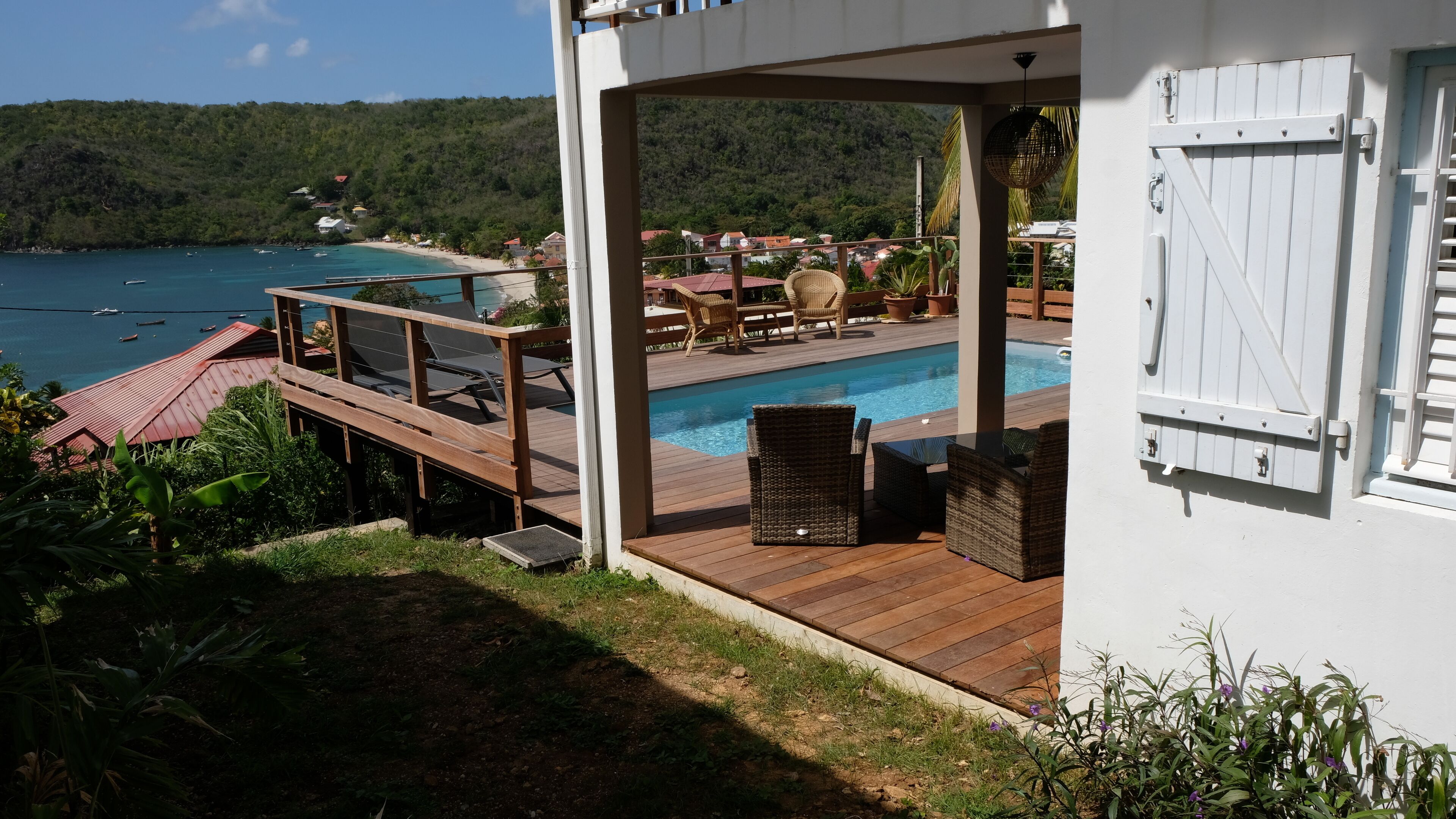Une terrasse en bois avec une piscine, des meubles en osier et une vue sur la mer et les collines.