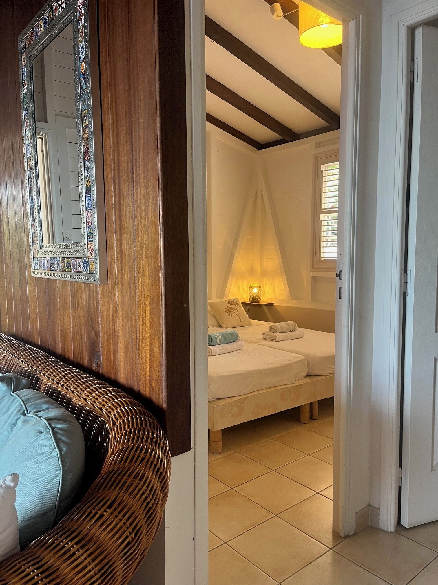 Een kamer met een rieten bank, een bed met witte lakens, een decoratieve spiegel en een warme lamp.