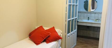 A small, narrow room with a bed and a bathroom visible through an arched doorway.