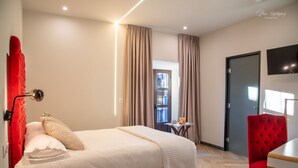 A hotel room with a bed, a red chair, a table with a glass of orange juice, and a TV on the wall. - Palacio de San Fernando (Utrera)