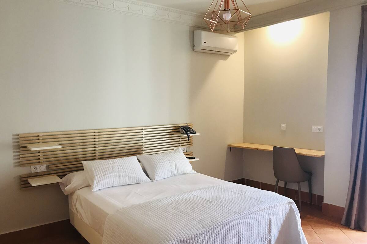 A bedroom with a wooden headboard, a bed with white bedding, a desk with a chair, and an air conditioning unit on the wall.