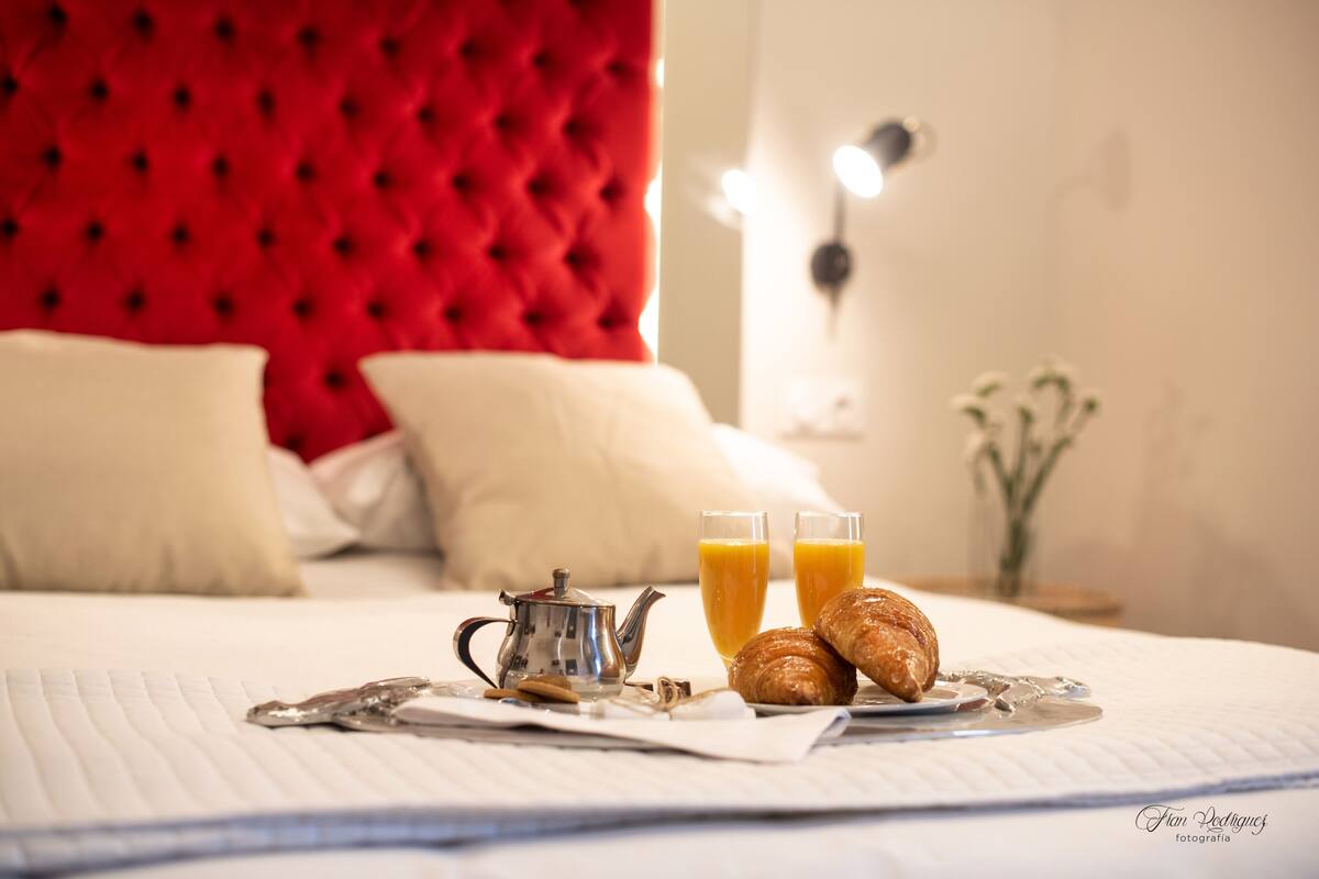 A neatly made bed with a red headboard, a teapot, two glasses of orange juice, and croissants on a white linen.