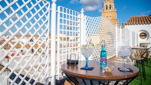 A table with two glasses, a bottle, and a bowl of fruit, set against a white lattice fence with a building in the background. - Palacio de San Fernando (Utrera)