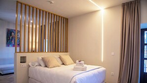 A hotel room with a bed, a wooden slat headboard, a bedside table, a wall-mounted light, and a window with curtains. - Palacio de San Fernando (Utrera)