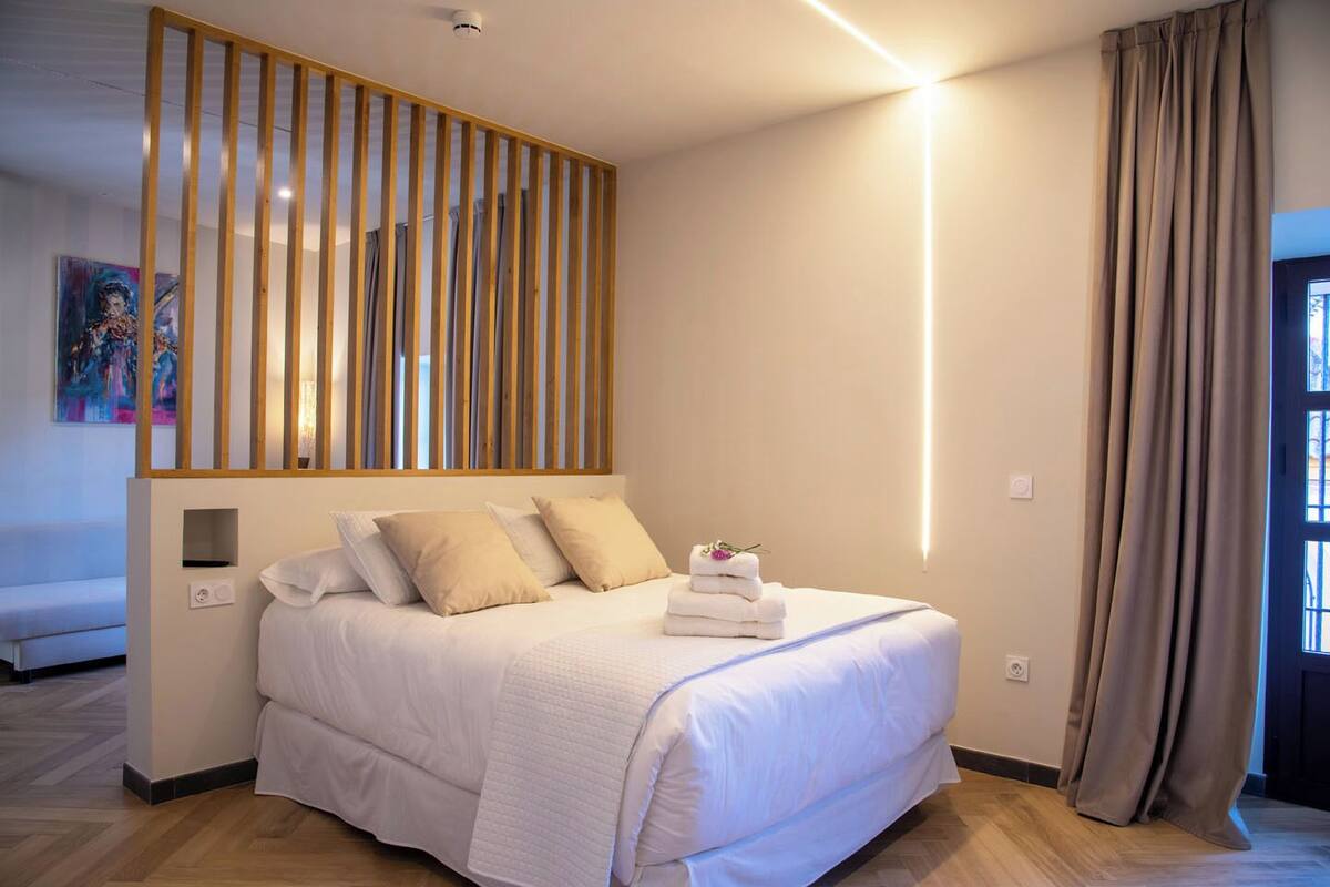 A hotel room with a bed, a wooden slat headboard, a bedside table, a wall-mounted light, and a window with curtains.