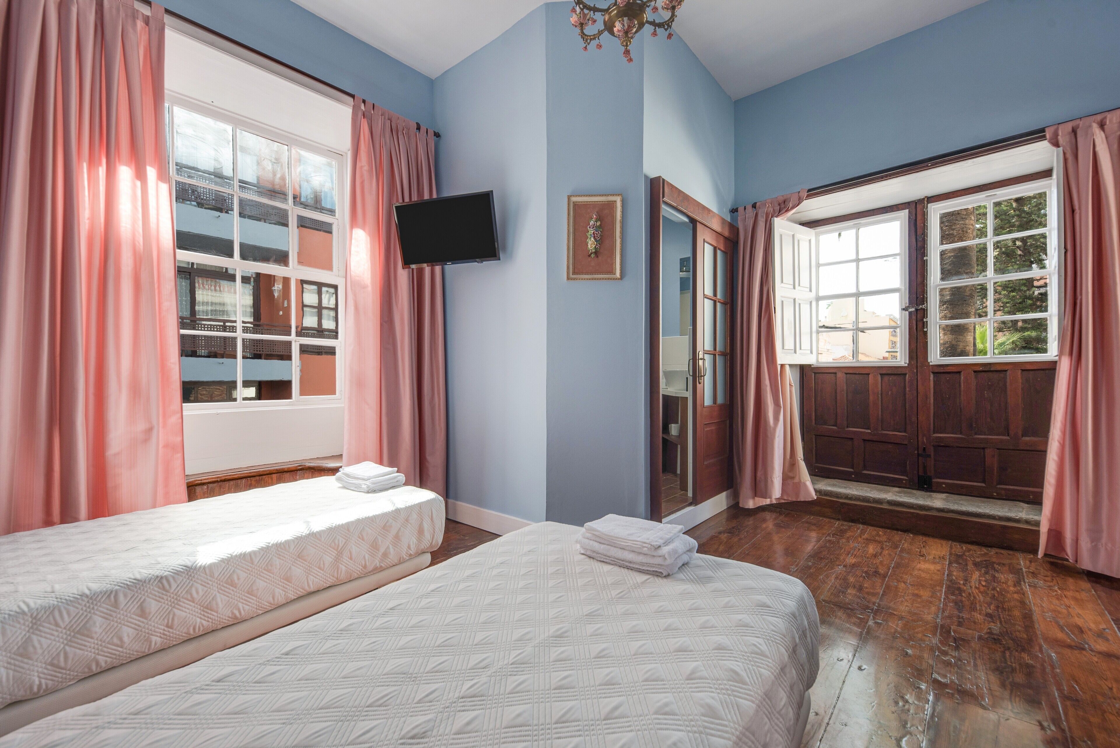 A bedroom with two beds, a television, a mirror, and a large window with curtains.
