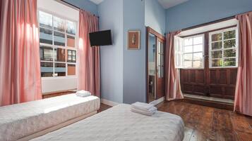A bedroom with two beds, a television, a mirror, and a large window with curtains.