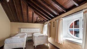 Een slaapkamer met twee enkele bedden, een groot raam met gordijnen, houten balken aan het plafond en een witte hoofdbord.