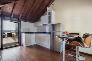 Une cuisine avec des meubles blancs, un réfrigérateur en acier inoxydable, une table à manger en bois avec deux tabourets de bar, et une vue sur un espace de vie comprenant un canapé et une petite table.
