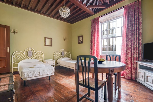 Une pièce dotée d’un plancher en bois, de deux lits simples avec du linge de lit blanc, d’une table à manger avec des chaises, d’un lustre et d’une fenêtre avec des rideaux.