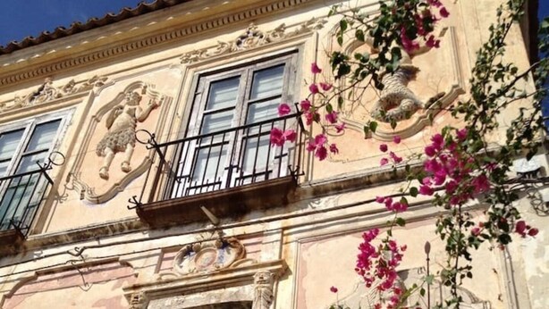 A building facade with classical architectural elements, including a balcony with a wrought iron railing, a window with green shutters, and decorative sculptures. - Serenity Escape in Marina Di Caulonia (Caulonia)