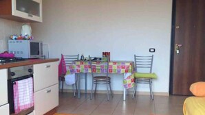 A small kitchen with a microwave, stove, and oven. There is a dining table with chairs and a colorful tablecloth. A door is visible on the right side. - Serenity Escape in Marina Di Caulonia (Caulonia)