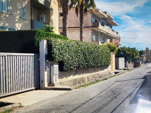 A residential street with a gated entrance, a hedge, and a multi-story building with balconies.