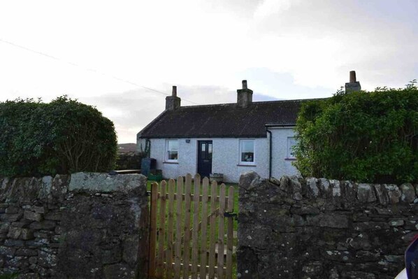 A white cottage with a black door, surrounded by stone walls and a wooden gate.