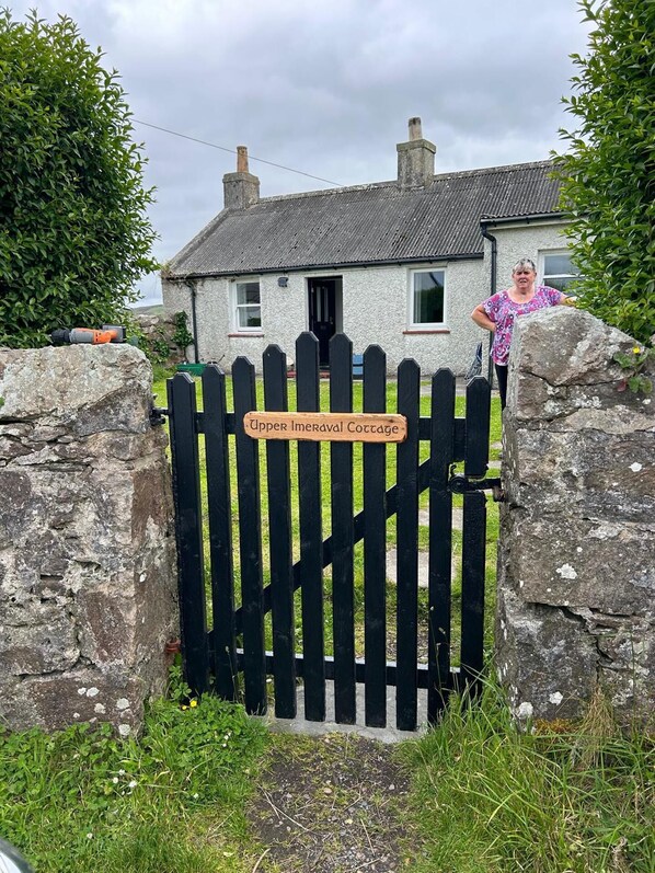 A cottage with a black picket gate, a stone wall, and a sign reading 'Upper Imeraval Cottage.'