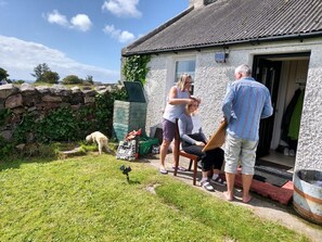 A stone building with a metal roof, a garden with a stone wall and a dog, and a person painting.