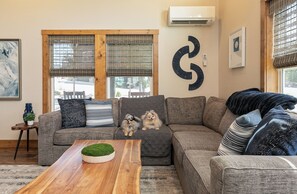 Una sala de estar con un sofá seccional, dos perros pequeños, una mesa de centro de madera y un aire acondicionado instalado en la pared.