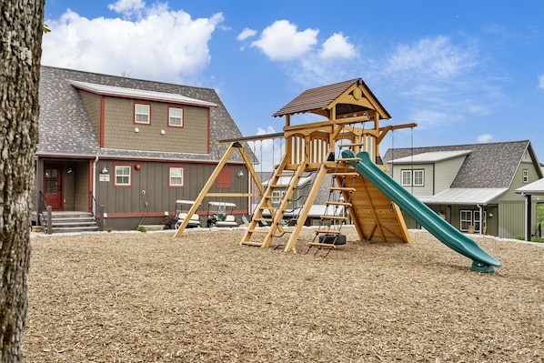 A playground with a wooden slide and climbing structure, surrounded by a wooden mulch ground cover, with houses in the background.