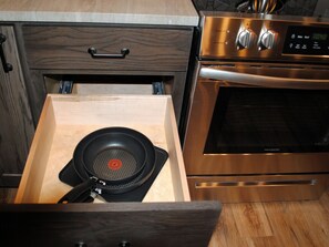 A kitchen with a drawer containing a frying pan, a stainless steel oven, and a wooden cabinet with a handle.
