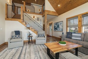 A spacious living area with a wooden staircase, two sofas, a coffee table, and a large window with blinds.