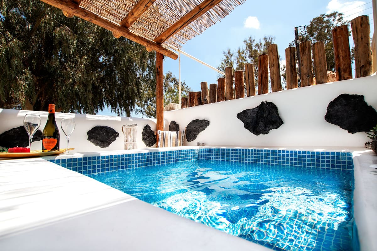 A swimming pool with blue tiles, a wooden pergola, and a table with wine bottles and glasses.