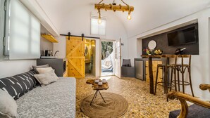 Un interior acogedor con un sofá, un tapete redondo, una mesa de madera con taburetes, una lámpara colgante y una zona de cocina con un televisor.