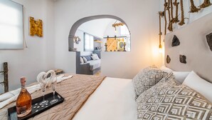 Un dormitorio con cama, una mesita de noche con una botella y vasos, una colcha estampada, y una vista a través de un arco hacia otra habitación.