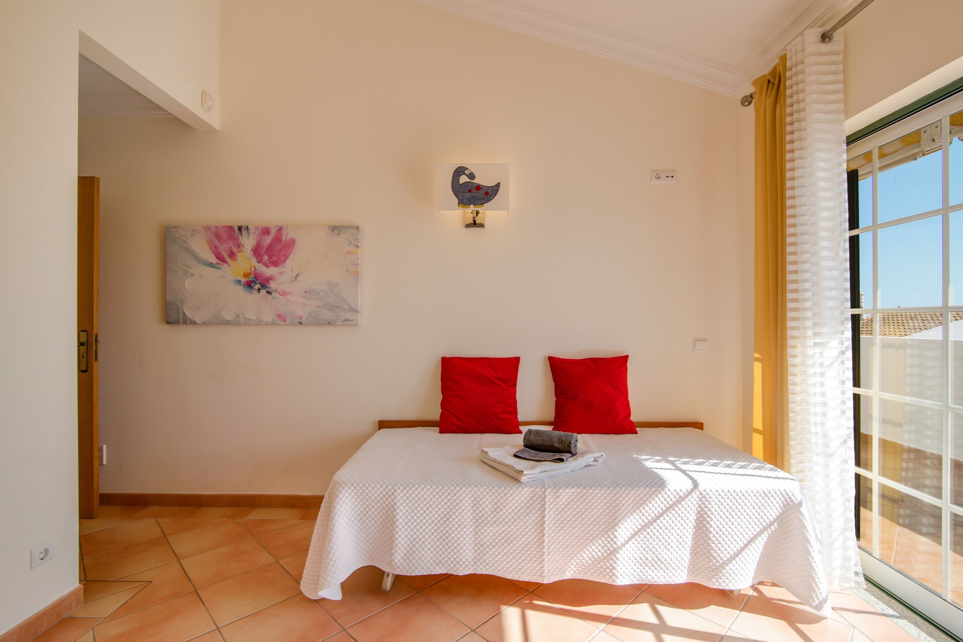 A bedroom with a bed, red pillows, a painting, and a window with curtains.