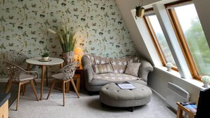 A cozy room with a round table, a sofa, and a chair. There is a window with a view of greenery outside. - Serendipity- Romantic 1-bed Apt Ambleside (Ambleside)