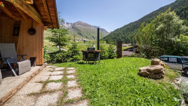 Una baita di legno con camino in pietra, un vialetto sterrato e vista su una montagna.