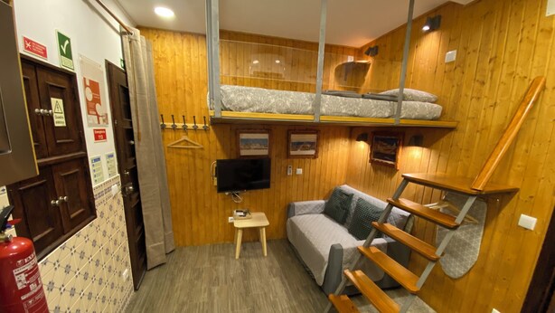 A compact room with a bunk bed, a sofa, a small table, and a TV mounted on the wall. There are wooden walls and a fire extinguisher on the left. - Estudio T34 - City Historic Center (Lisboa)