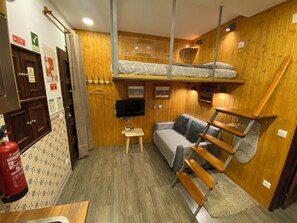 Une petite pièce avec un lit superposé, un canapé, une petite table et un téléviseur fixé au mur. Les murs sont en bois et il y a un extincteur sur le côté gauche.