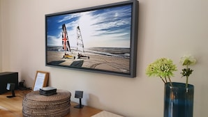 A framed painting of a sailboat on a beach, a small table with a speaker, and a vase with flowers on the wall.