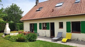 Une maison avec un toit recouvert de tuiles, des volets verts et une cheminée. On y trouve une terrasse avec une table et des chaises, ainsi qu’une chaise jaune à l’extérieur.