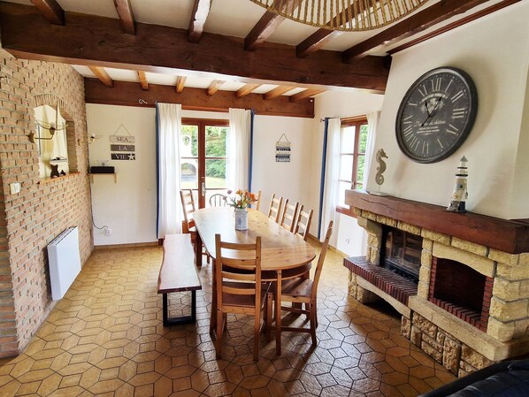 A dining area with a wooden table, chairs, a bench, a fireplace, and a large wall clock.