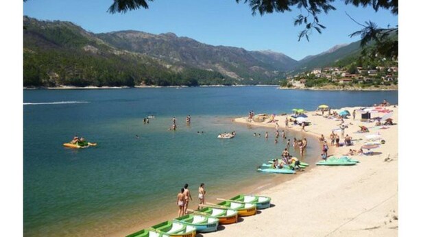Une plage bordée de bateaux, où l’on peut voir des gens se baigner, avec une chaîne de montagnes en arrière-plan.