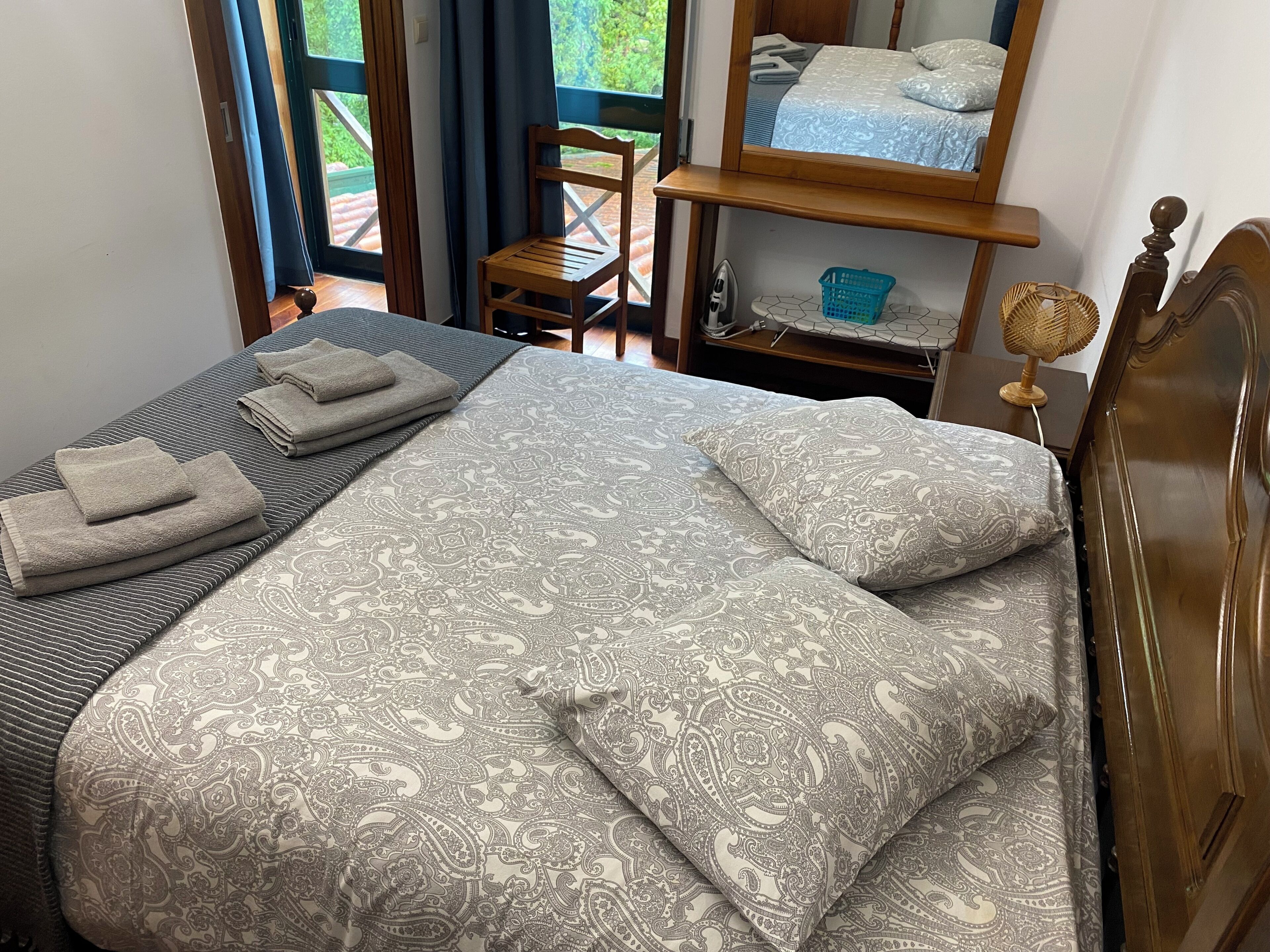 Una cama bien hecha con una colcha estampada y almohadas, una silla de madera, una mesita con una cesta y un espejo que refleja parte de la habitación.