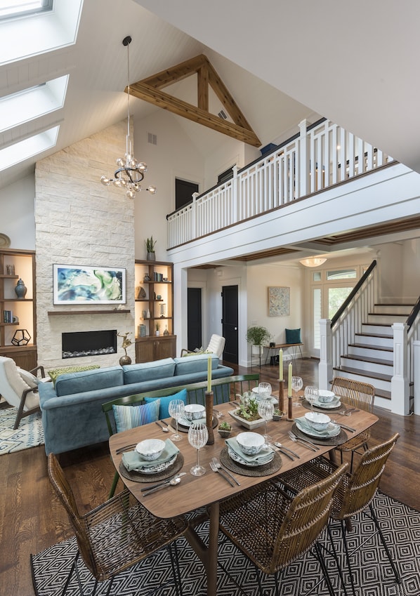 A spacious living area with a high ceiling, a fireplace, a dining table set for six, and a staircase leading to an upper level.