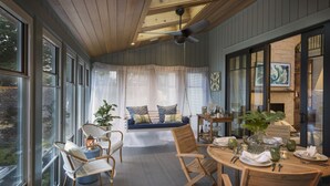A cozy dining area with a round table set for four, a swing seat with cushions, and large windows offering a view of the outdoors. - The "Idea House" From TV Series This Old House! High End, Definition of Luxury! (Narragansett)