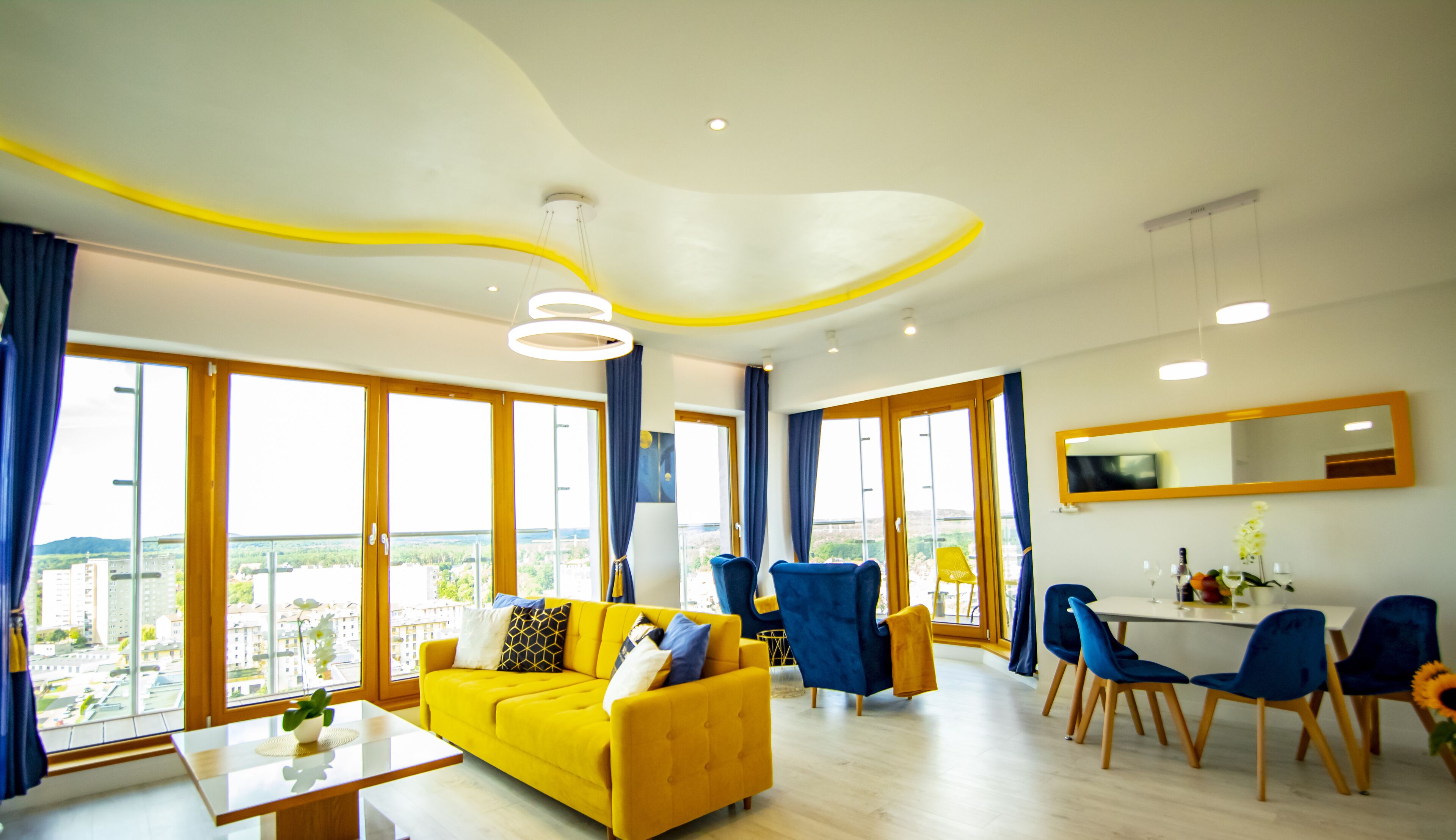 A modern living room with a yellow sofa, blue chairs, and a dining area with a white table and blue chairs.
