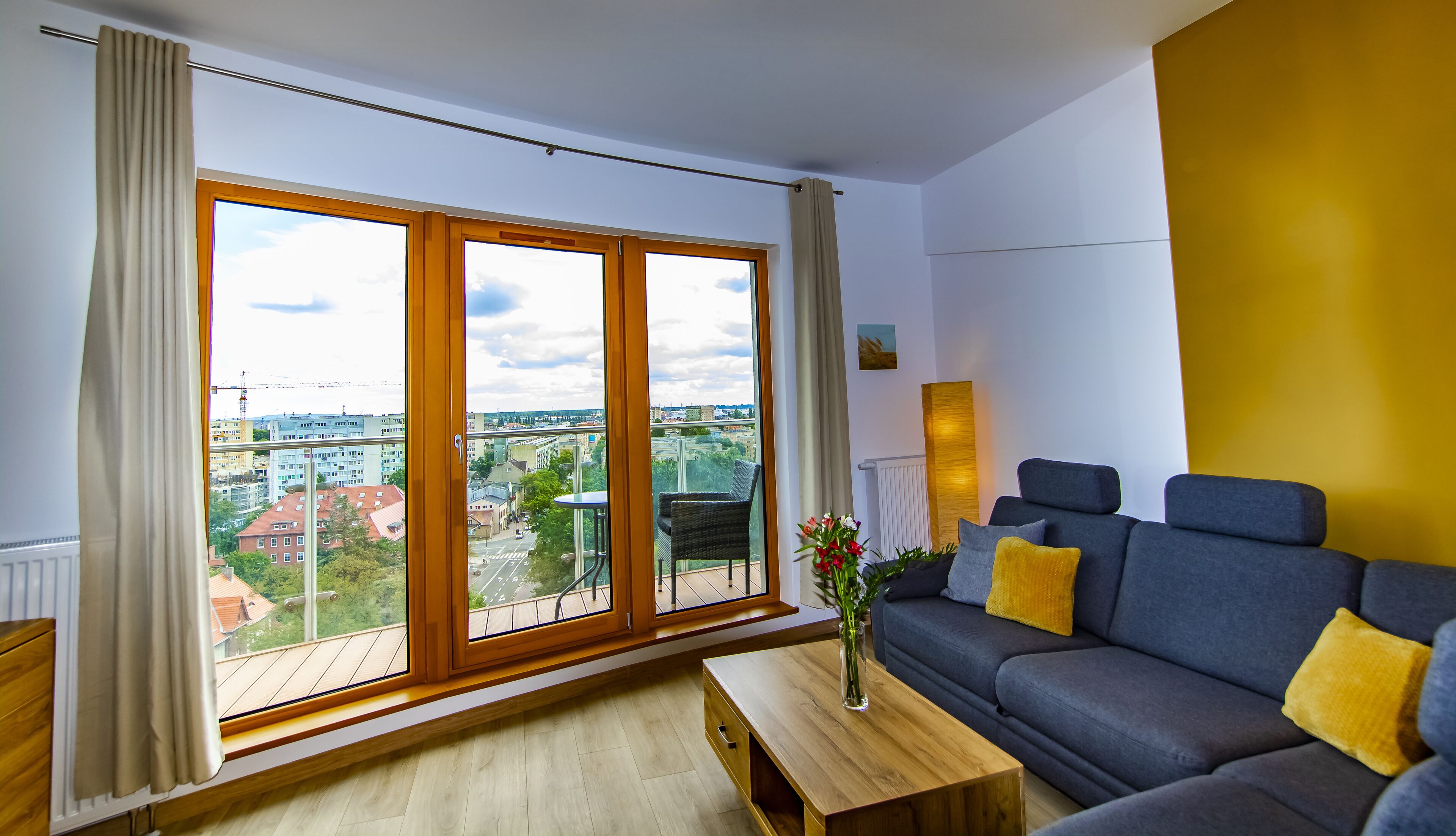 Ein modernes Wohnzimmer mit großem Fenster, einem grauen Sofa mit gelben Kissen, einem Holztisch und einer Vase mit Blumen.