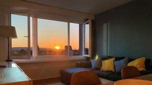 A modern living room with a large window overlooking a cityscape at sunset. - Lake View Business Apartment (Zug)
