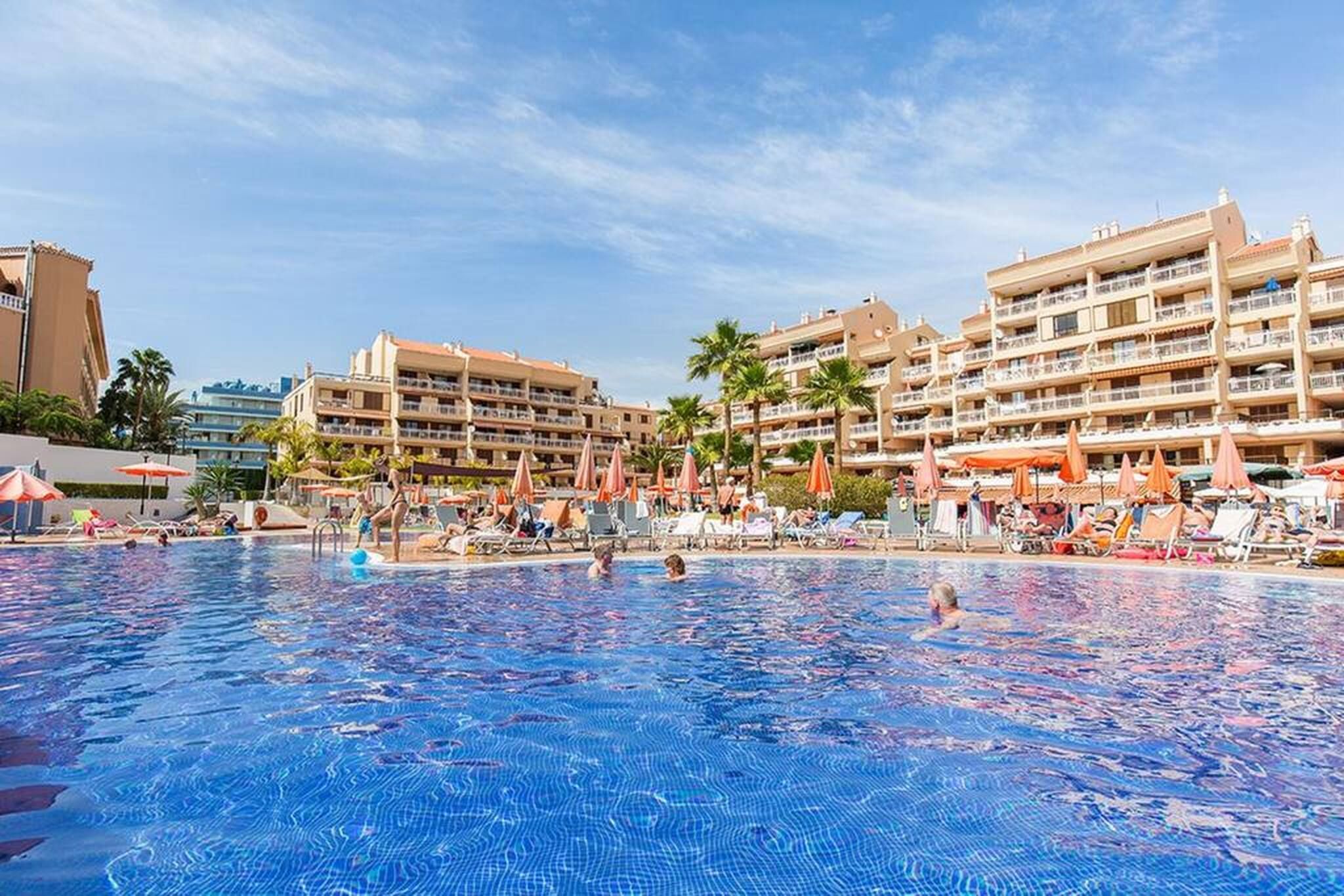 Una piscina rodeada de tumbonas y sombrillas, con edificios de varios pisos al fondo.