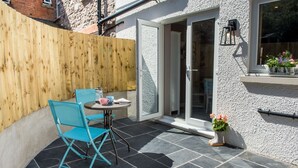 A patio with a table and chairs, a wooden fence, and a building with a white exterior and a window with flowers. - Perfect romantic break-  cottage in Laugharne (Laugharne)
