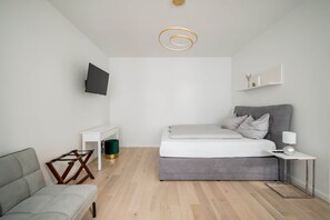 A modern bedroom with a grey sofa, a bed with grey bedding, a small white bedside table, a wall-mounted TV, and a ceiling light fixture. - Prime Apartment, Prime Apartment nähe Naschmarkt (Vienna)