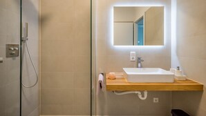 Un baño moderno con cabina de ducha de vidrio, tocador de madera con lavamanos blanco y un espejo grande con iluminación integrada.